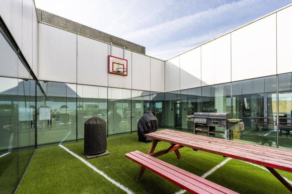 Rooftop terrace on Krijn Taconiskade with artificial grass, red picnic tables, a basketball hoop, and a barbecue grill.